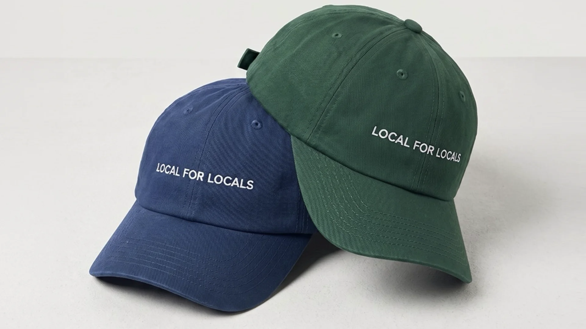 Dois bonés de estilo "dad hat" sobrepostos em um fundo cinza claro. O boné de baixo é azul-marinho e o de cima é verde-escuro. Ambos possuem a frase "LOCAL FOR LOCALS" bordada em linha branca na parte frontal.