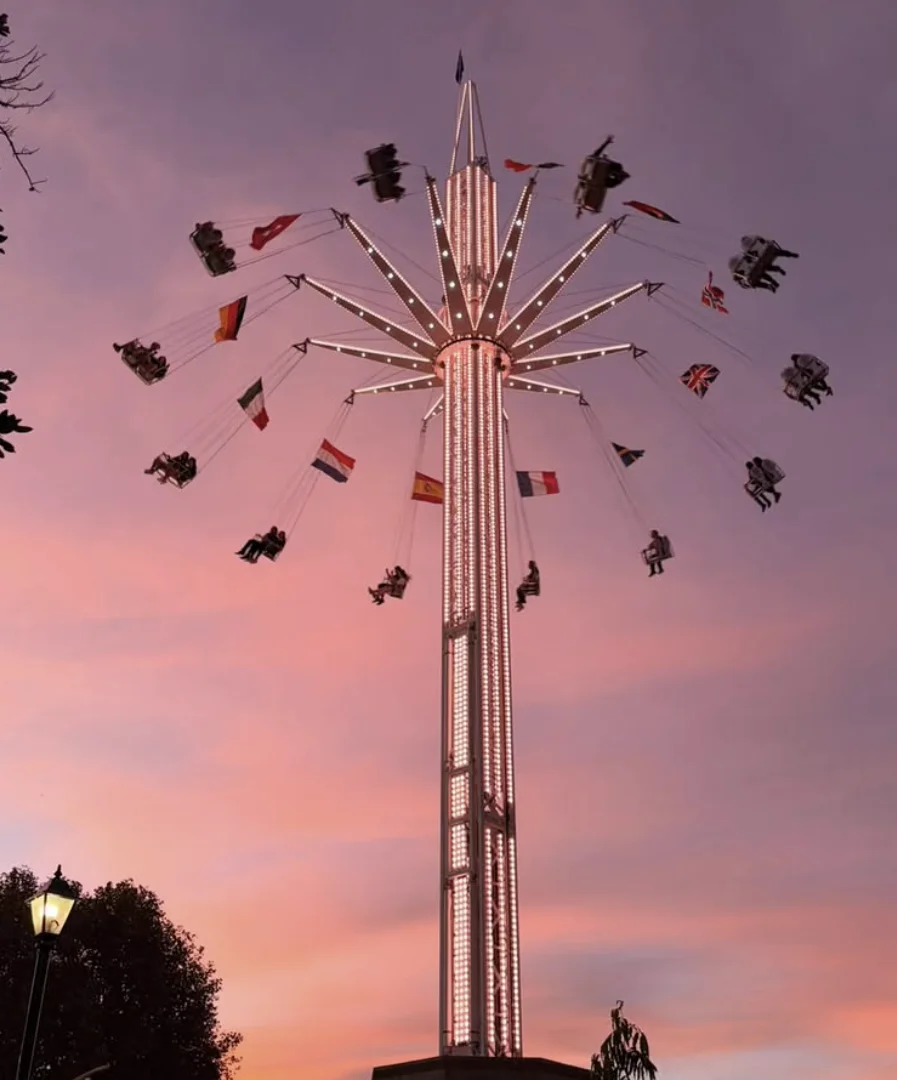 Brinquedo do tipo torre de cadeiras voadoras giratórias à noite, todo iluminado, com pessoas nas cadeiras e bandeiras de vários países penduradas. O fundo exibe um céu de fim de tarde com tons de rosa e roxo.