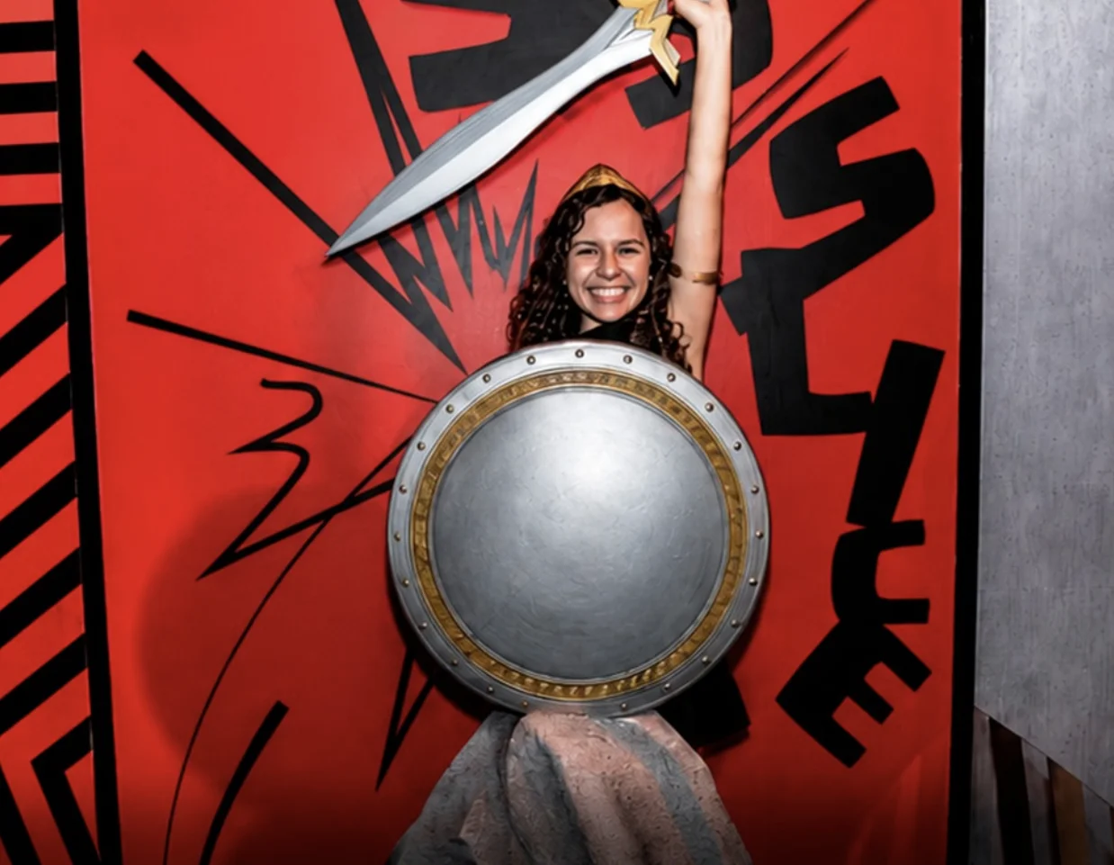 Jovem sorridente fantasiada de Mulher-Maravilha segurando espada e escudo em cenário vermelho.