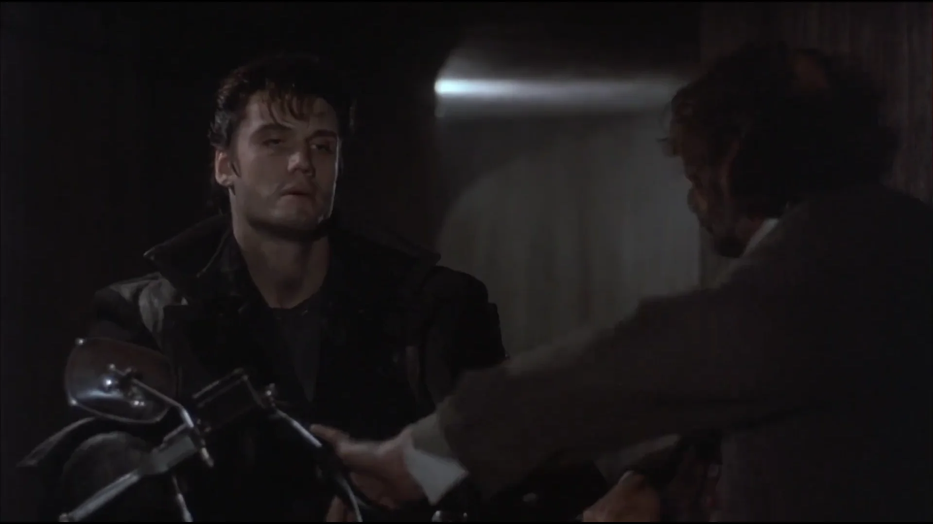 Frank Castle, interpretado por Dolph Lundgren, sentado em uma motocicleta em um ambiente escuro, conversando com um homem de costas.