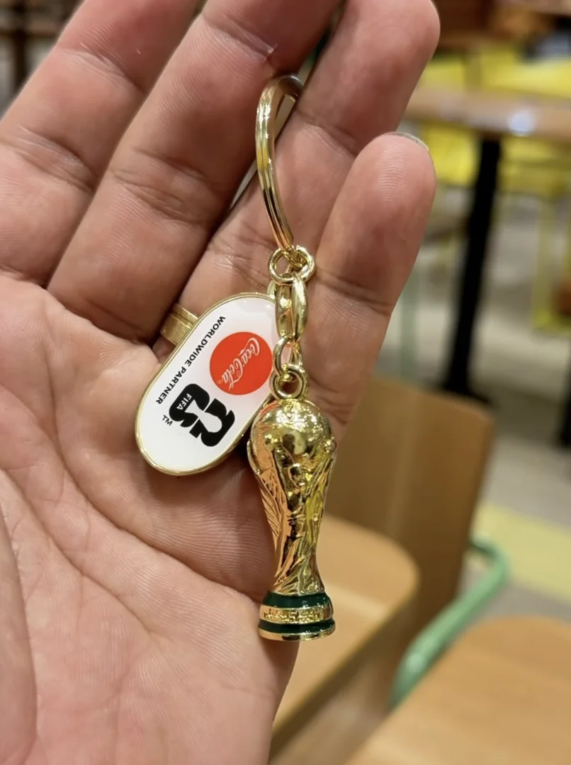 Mão segurando um chaveiro dourado com a réplica em miniatura da Taça da Copa do Mundo da FIFA e uma placa oval com os logos da Coca-Cola e da Copa de 2026.