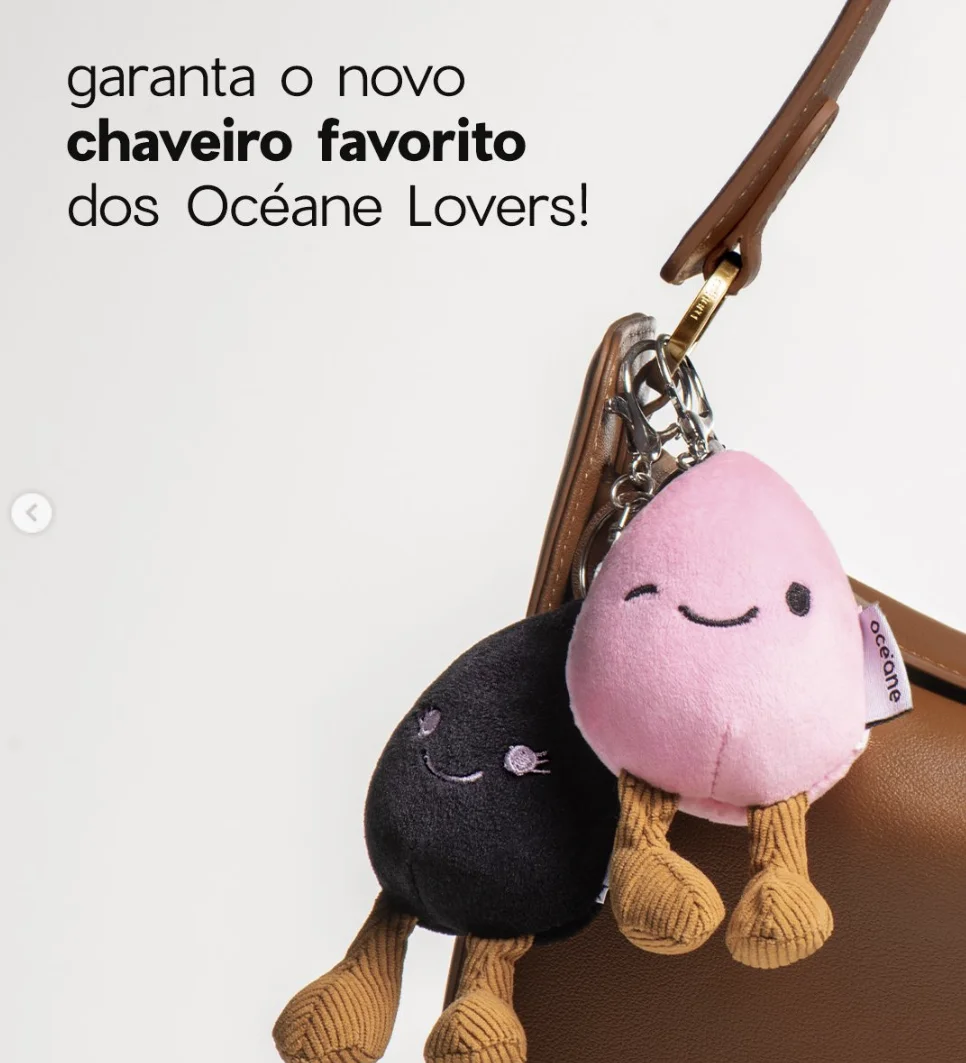 Dois chaveiros de pelúcia em formato de esponja de maquiagem, um rosa e um preto, pendurados em uma bolsa de couro marrom. Eles têm rostos sorridentes e perninhas de veludo cotelê.
