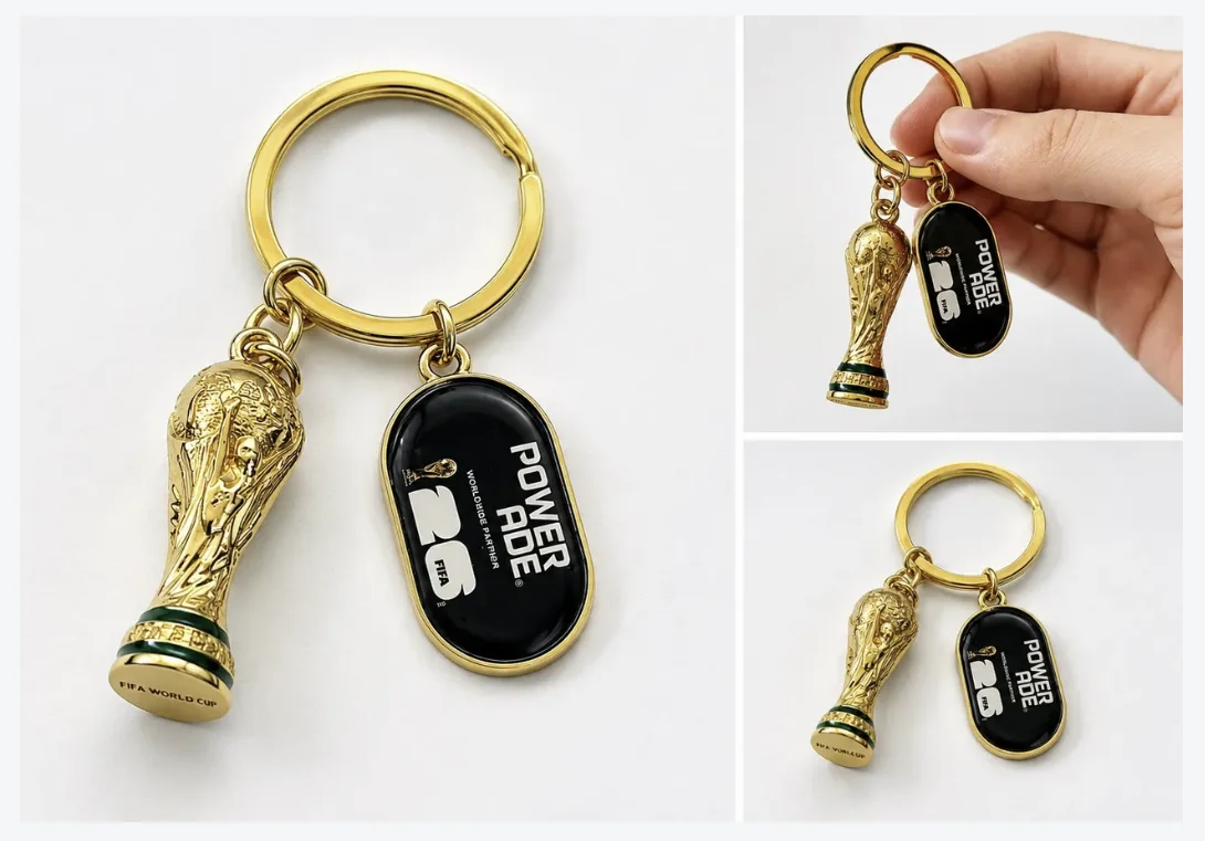 Close-up e detalhes de um chaveiro dourado com a miniatura da Taça do Mundo da FIFA e uma placa preta com o logotipo da Powerade e da Copa de 26.