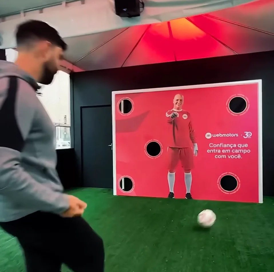 Homem de perfil chutando uma bola de futebol em direção a um painel vermelho com alvos em um espaço interno.