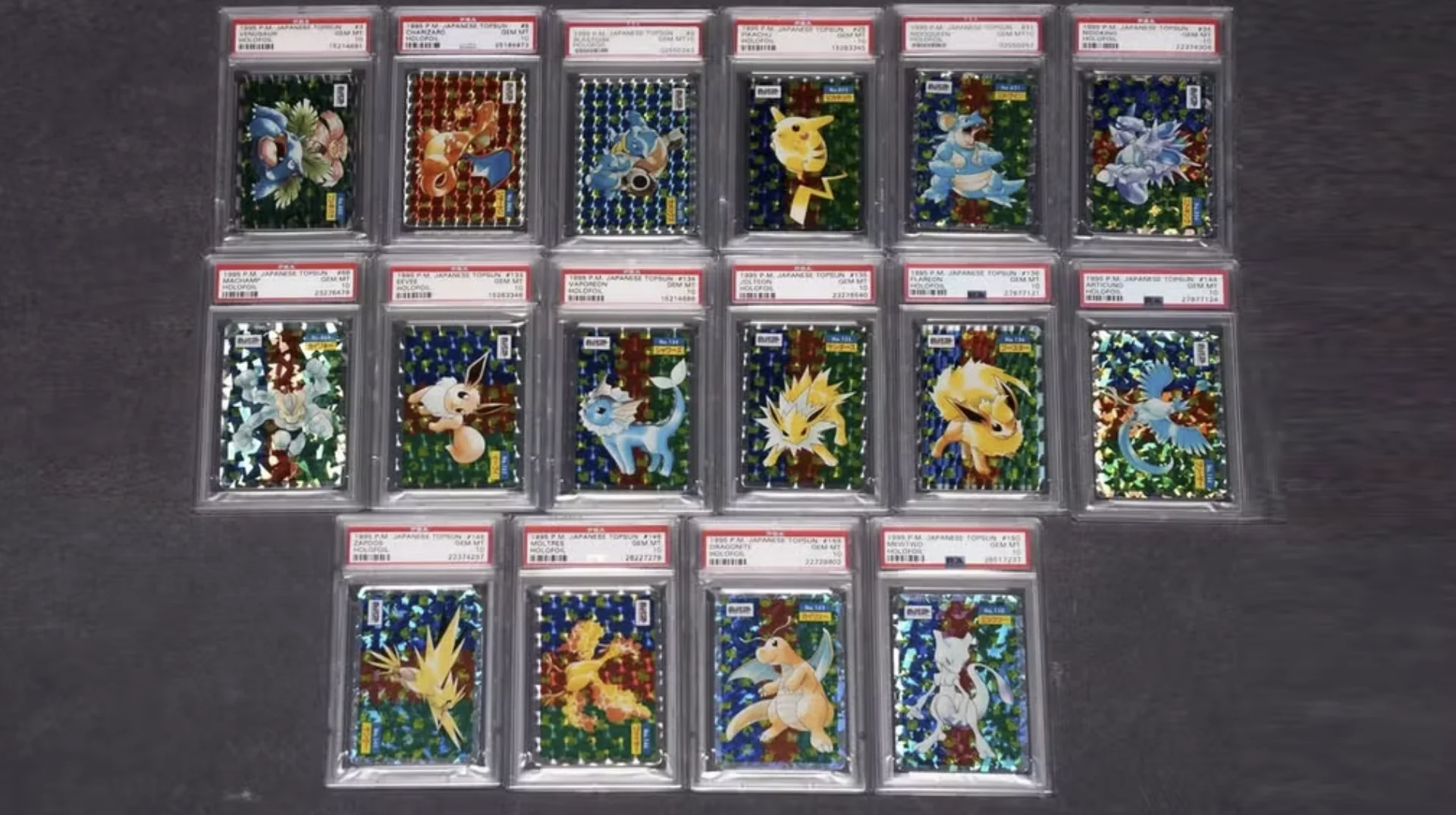 Dezesseis cartas Pokémon japonesas antigas e brilhantes, todas protegidas em cases de acrílico da PSA com etiquetas vermelhas na parte superior, organizadas em três fileiras sobre um fundo cinza-escuro.