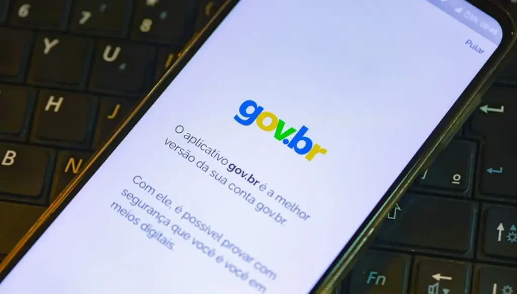 Celular apoiado sobre o teclado de um notebook, exibindo na tela o logotipo azul e amarelo do gov.br com um texto sobre a segurança do aplicativo.