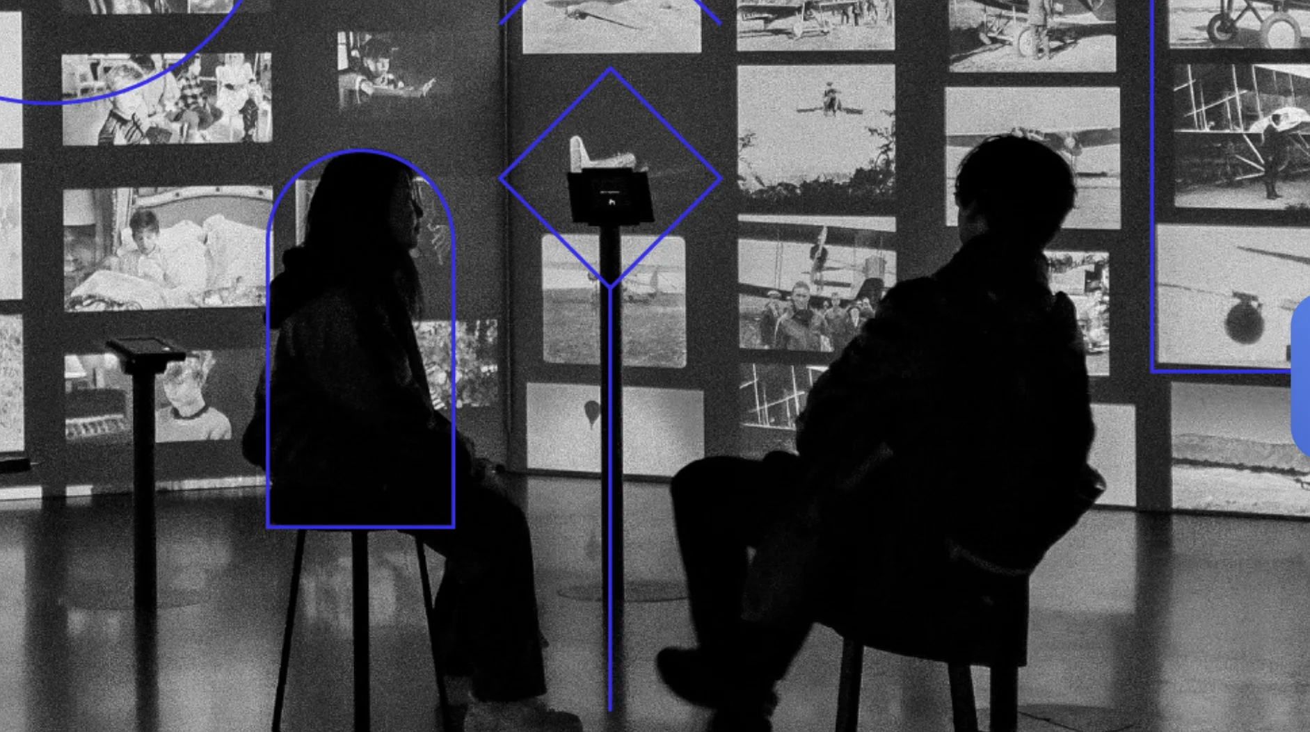 Duas pessoas sentadas de costas, em silhueta escura, observando uma grande parede repleta de fotografias históricas em preto e branco. Linhas geométricas azuis circulam e destacam partes da cena.