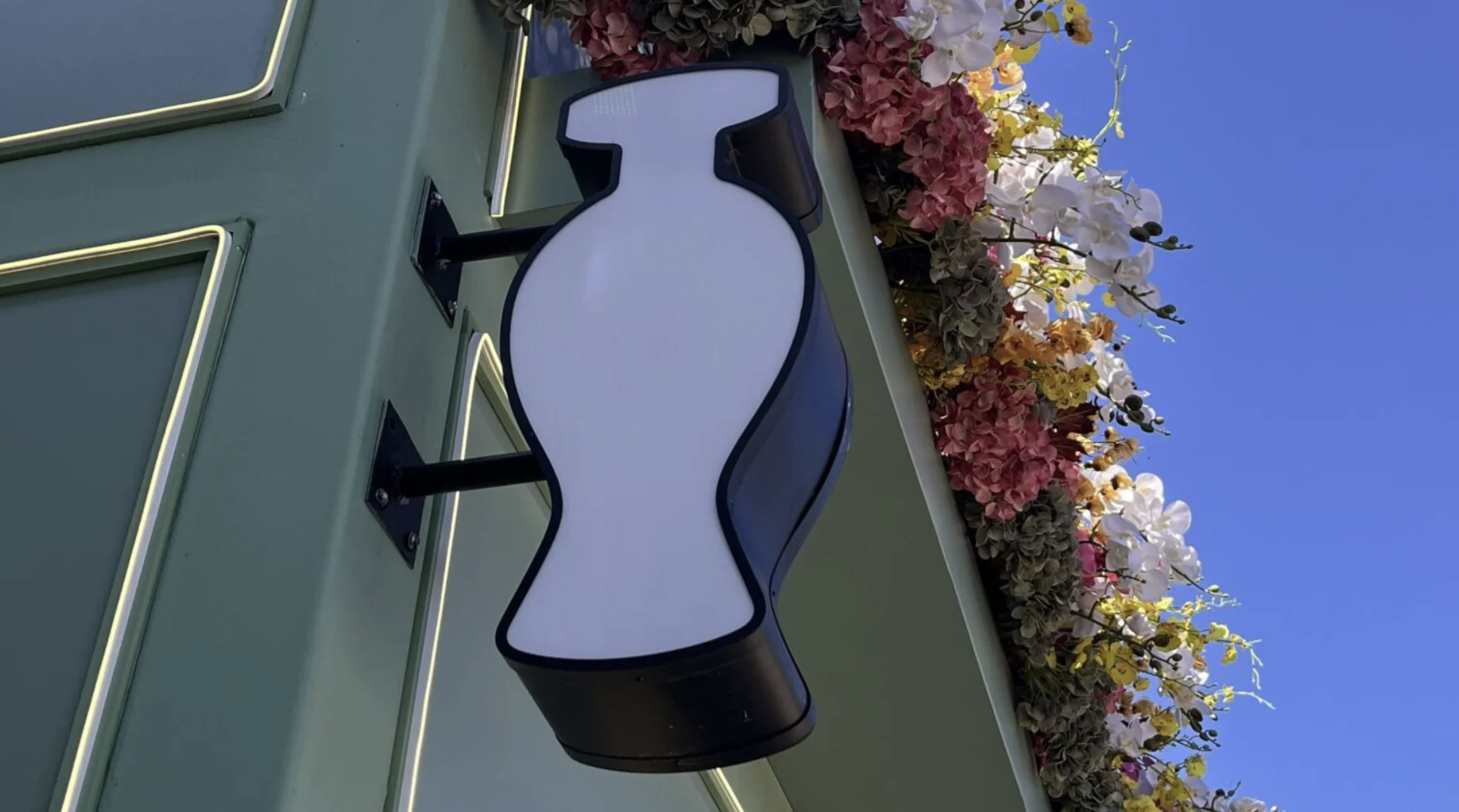 Um close-up de uma placa de loja com o formato de um frasco de perfume branco, montada em uma parede verde-oliva. No topo da foto, há um grande arranjo floral, e acima de tudo está um céu azul claro.