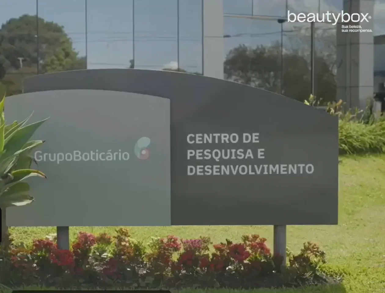 Placa externa cinza com a inscrição "GrupoBoticário Centro de Pesquisa e Desenvolvimento" em letras brancas, cercada por flores e gramado.