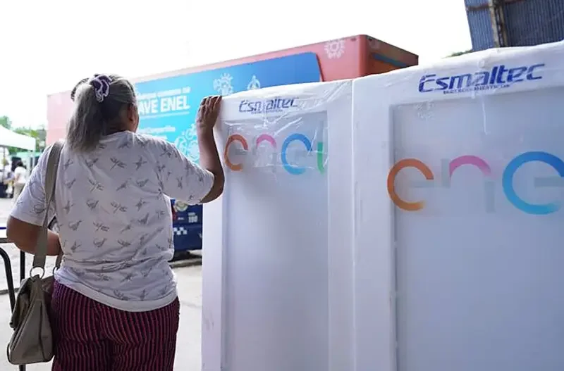 Mulher de costas, com camiseta branca e calça listrada, observa duas geladeiras novas da marca Esmalteca, ainda na embalagem de proteção com o logotipo da Enel.