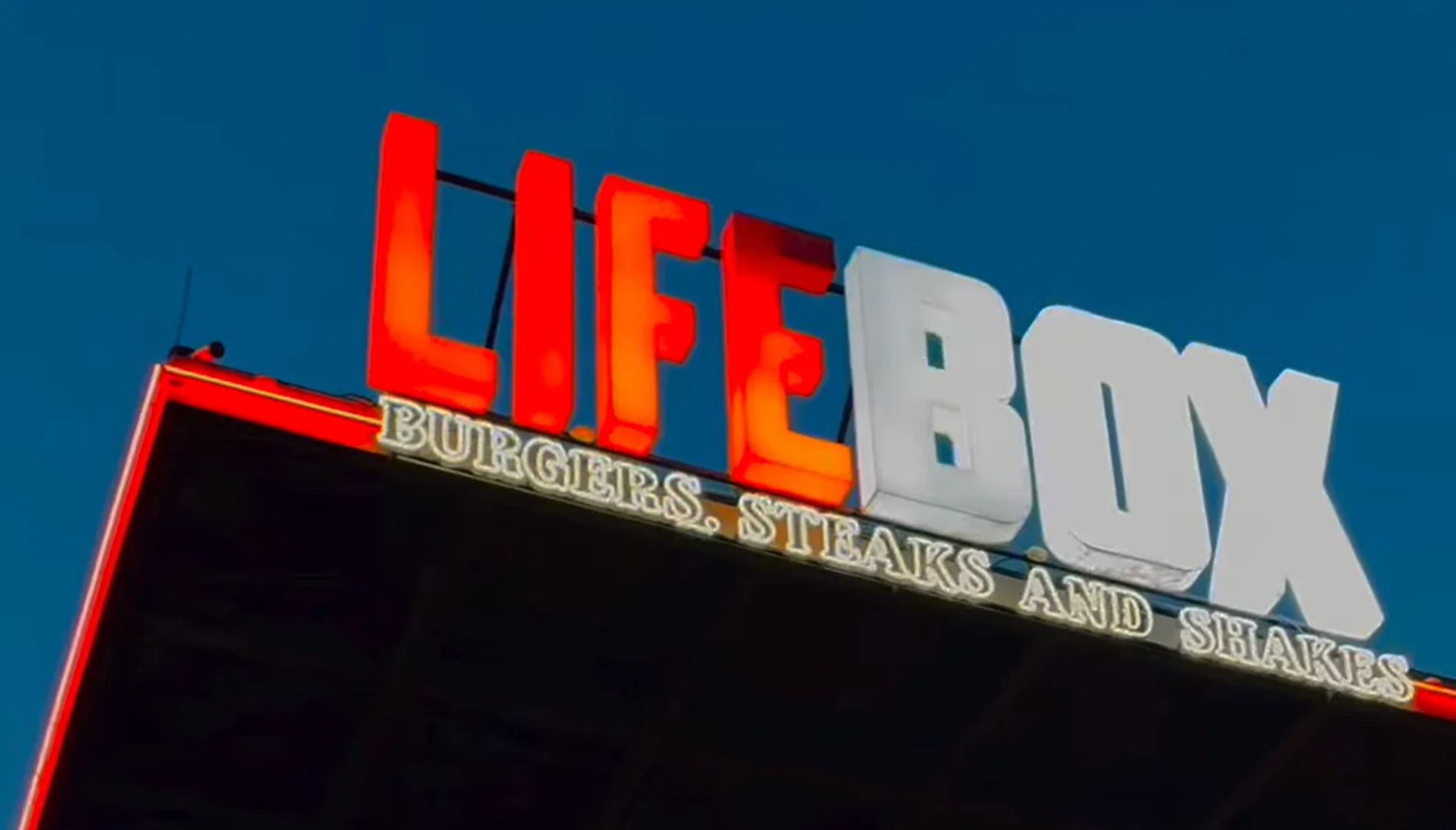 Letreiro luminoso de uma unidade da hamburgueria Lifebox contra um céu azul. A palavra "LIFE" está em letras garrafais vermelhas e iluminadas, e "BOX" em letras brancas. Logo abaixo, há a inscrição "BURGERS, STEAKS AND SHAKES" em letras menores e douradas.