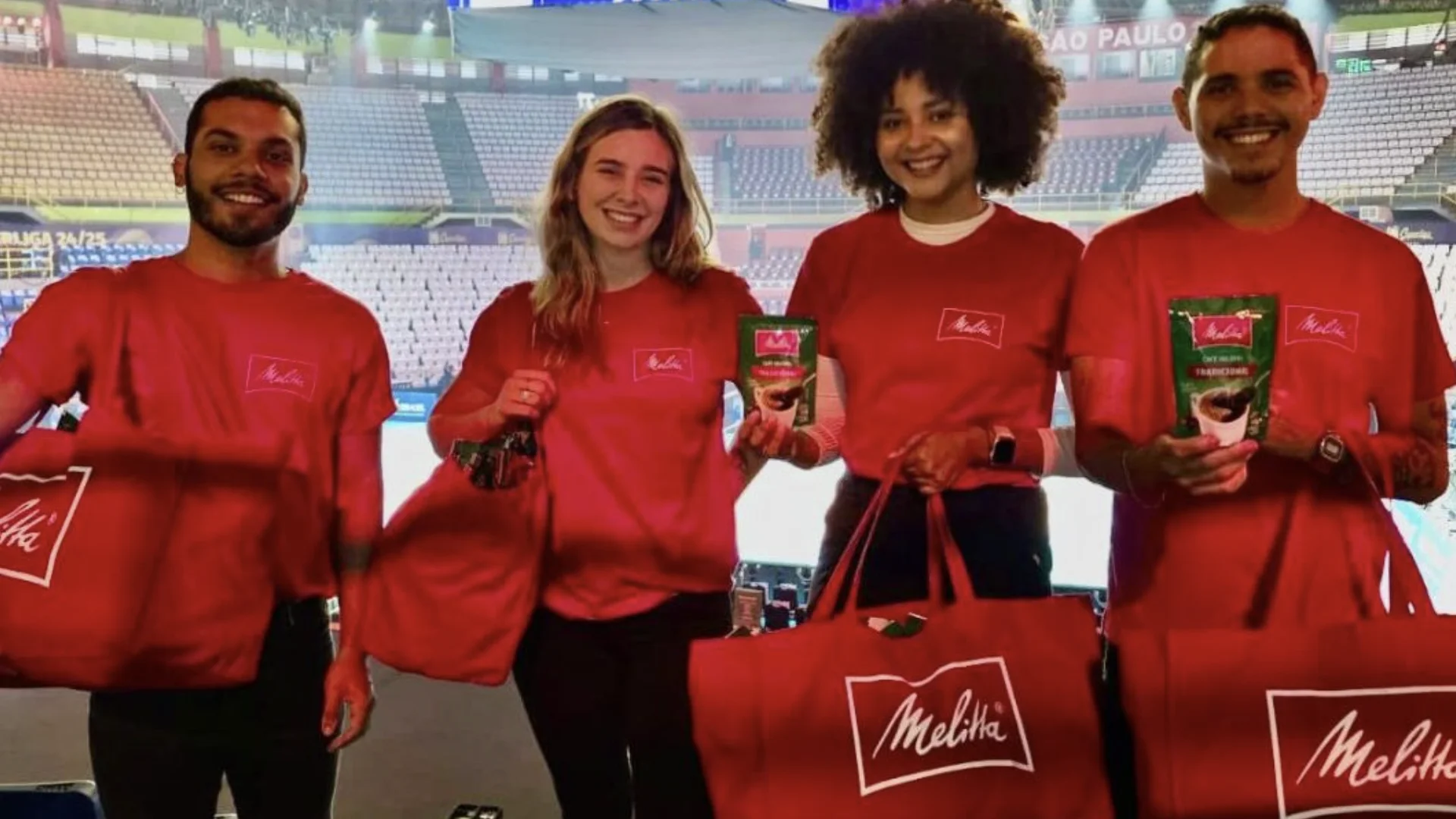 Quatro promotores da Melitta, vestindo camisetas vermelhas e segurando bolsas e embalagens de café, sorriem em um ginásio de esportes ao fundo.