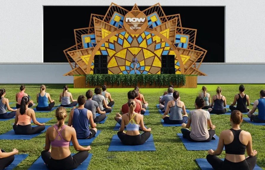 Vista ampla de uma aula de yoga ou meditação ao ar livre em frente ao palco decorado. Diversas pessoas estão sentadas de costas sobre tapetes azuis em um gramado verde, voltadas para uma instrutora no palco.