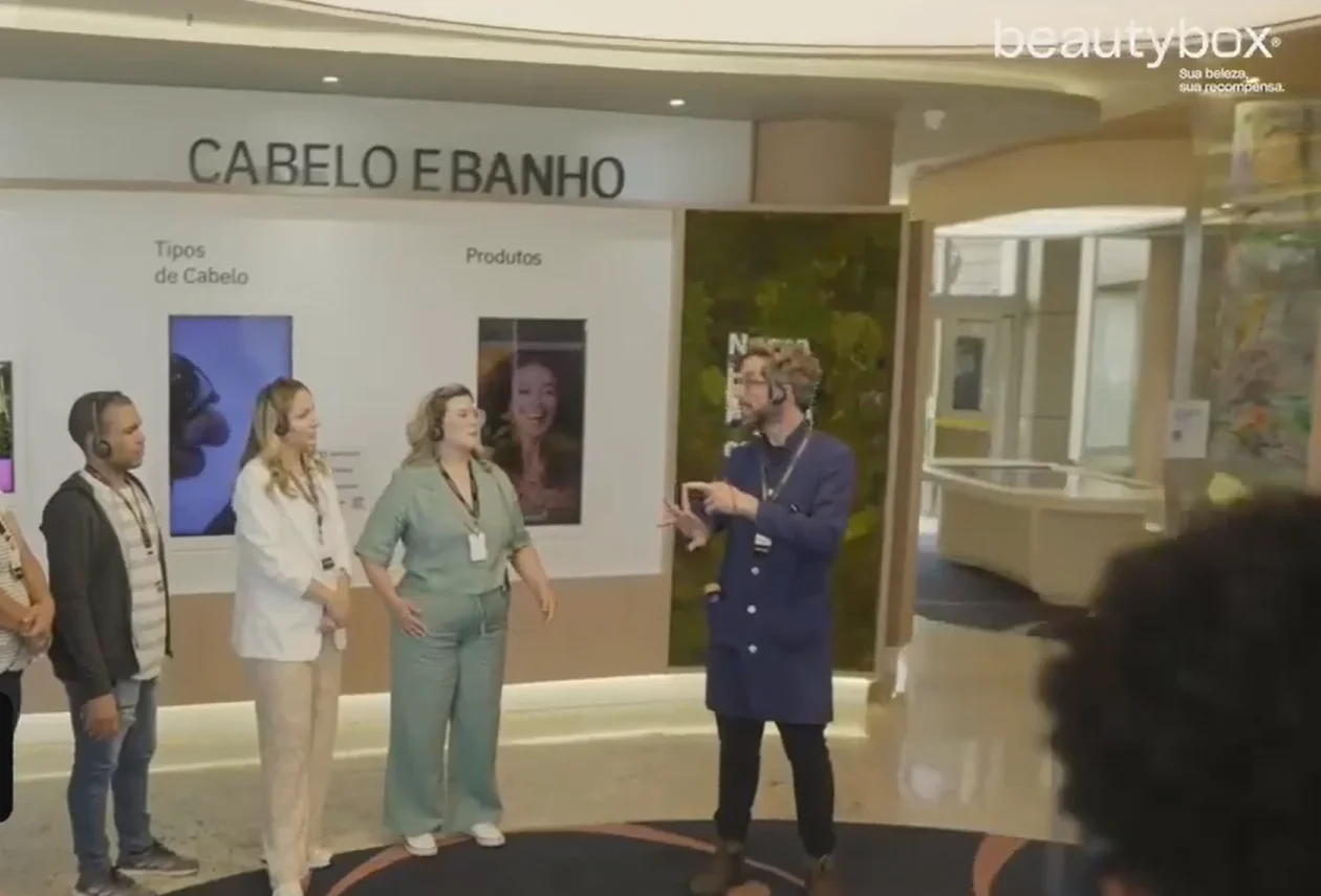 Homem em jaleco azul gesticula e explica algo para um grupo de visitantes em um painel interativo escrito "Cabelo e Banho".