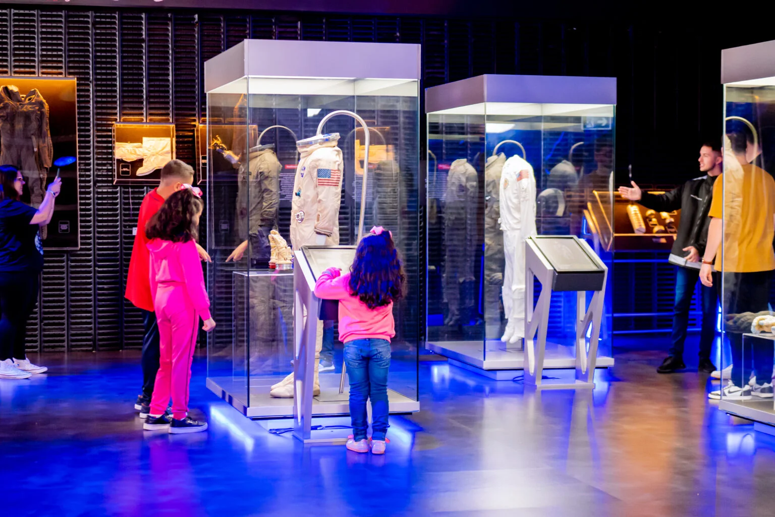 Crianças observam atentamente réplicas de trajes espaciais da NASA expostas em vitrines de vidro iluminadas em um ambiente de museu moderno.