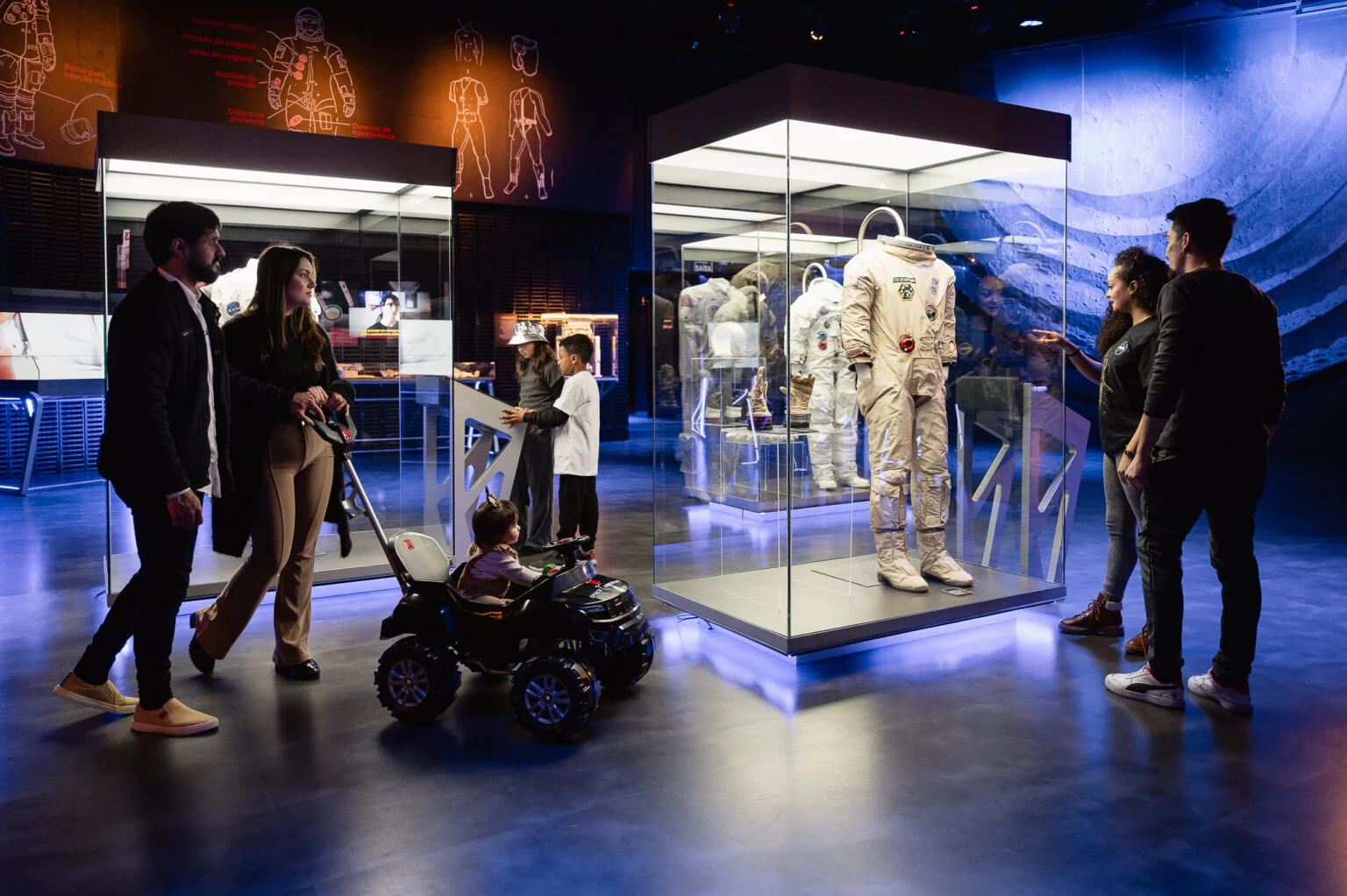 Uma família, incluindo um bebê em um carrinho de brinquedo, observa um traje de astronauta completo dentro de uma vitrine iluminada. Ao fundo, diagramas técnicos de naves espaciais.