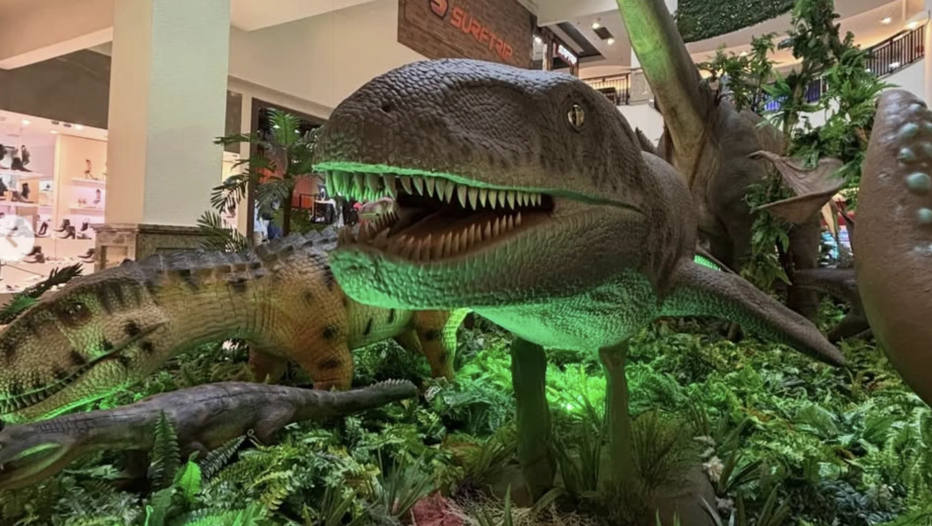 Close-up lateral de uma réplica de dinossauro aquático ou semiaquático com a boca aberta, revelando dentes afiados. A iluminação verde destaca a parte inferior da criatura em meio a plantas artificiais.