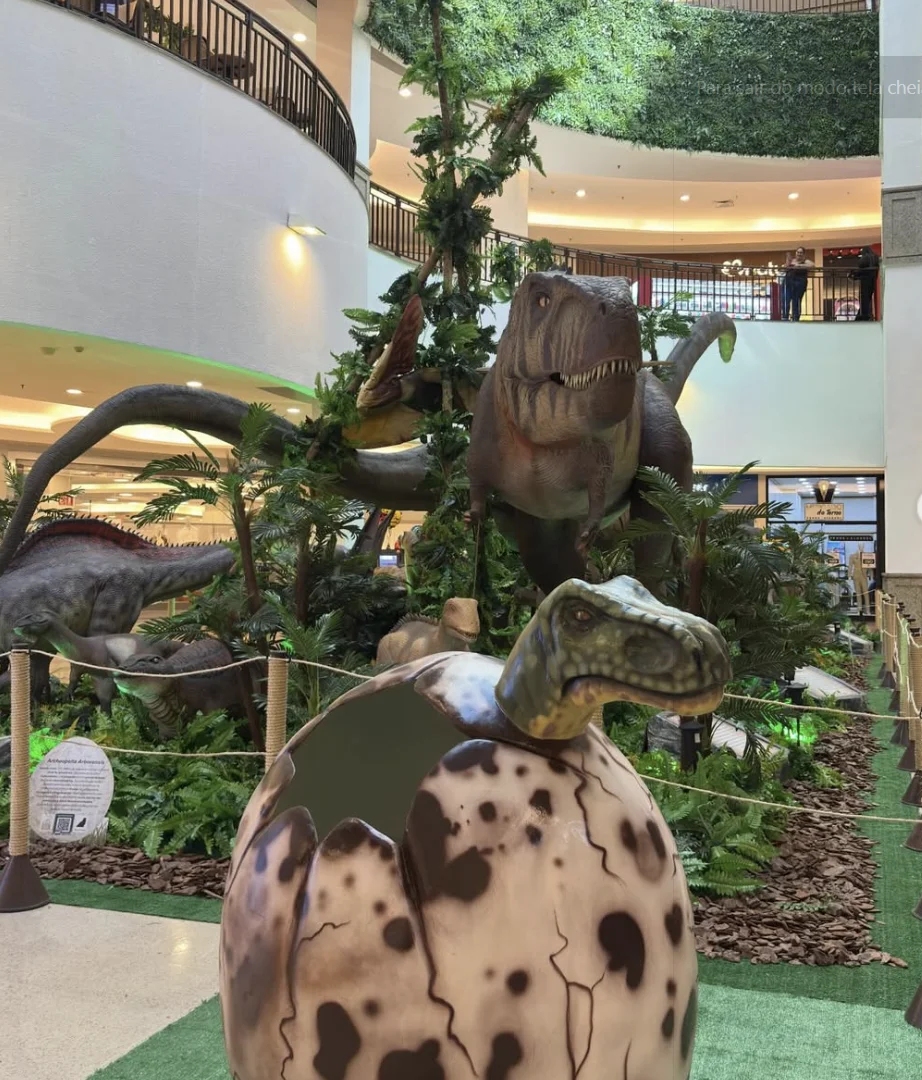 Em primeiro plano, um ovo de dinossauro gigante quebrado com um pequeno dinossauro saindo de dentro. Ao fundo, a mesma exposição com grandes dinossauros e o interior do shopping com vários andares.