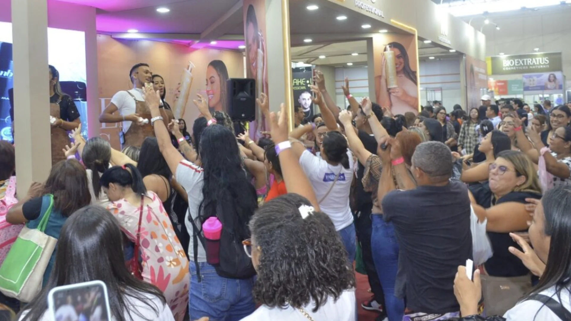 Multidão animada com braços levantados em frente a um palco de exposição de cosméticos em uma feira de beleza.