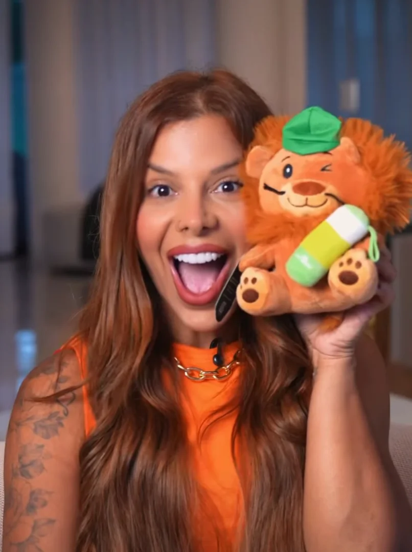 Uma mulher sorridente de cabelos ruivos longos segura e mostra uma pequena pelúcia de leão com chapéu verde e uma garrafinha térmica nas mãos.