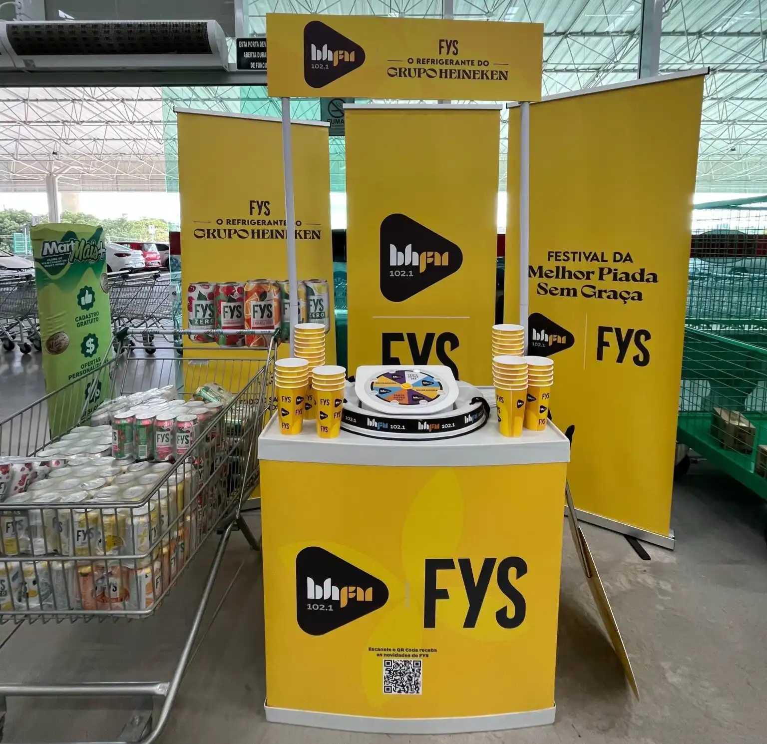 Estande promocional amarelo da marca de refrigerantes FYS e da rádio BH FM dentro de um supermercado, com um carrinho cheio de latas do produto ao lado.
