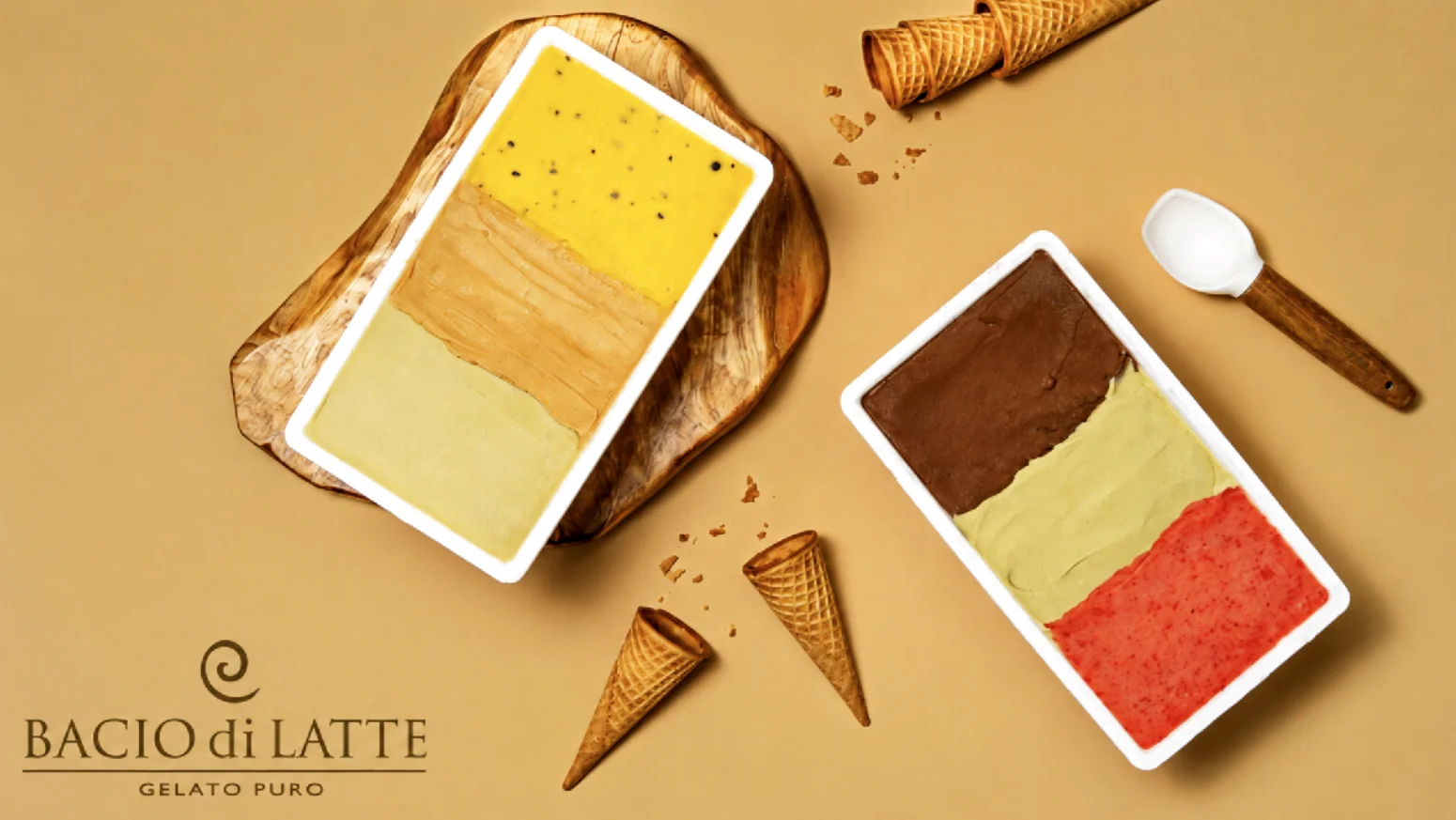 Foto de estúdio com fundo bege mostrando dois potes retangulares de gelato da Bacio di Latte. O pote da esquerda está sobre uma tábua de madeira rústica e traz três sabores: maracujá, doce de leite e pistache. O pote da direita contém os sabores chocolate, pistache e morango. Ao redor, há casquinhas de sorvete vazias, farelos e uma colher de pegar sorvete. O logotipo da marca está no canto inferior esquerdo.