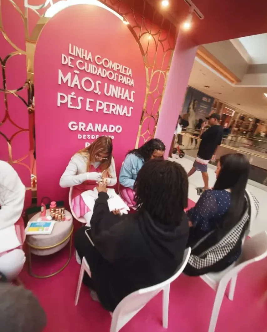 Área de manicure no quiosque da Granado. Clientes estão sentadas em cadeiras brancas recebendo serviços de esmaltação e cuidados com as unhas por profissionais. Ao fundo, um painel rosa diz "Linha completa de cuidados para mãos, unhas, pés e pernas".