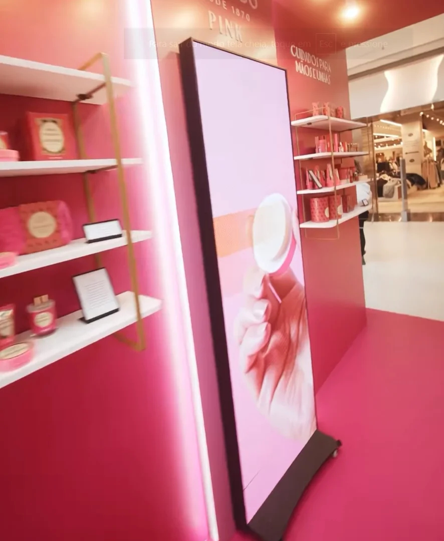 Interior do quiosque rosa exibindo prateleiras com produtos à esquerda e uma grande tela vertical digital ao centro, mostrando um vídeo promocional de mãos usando os cremes.