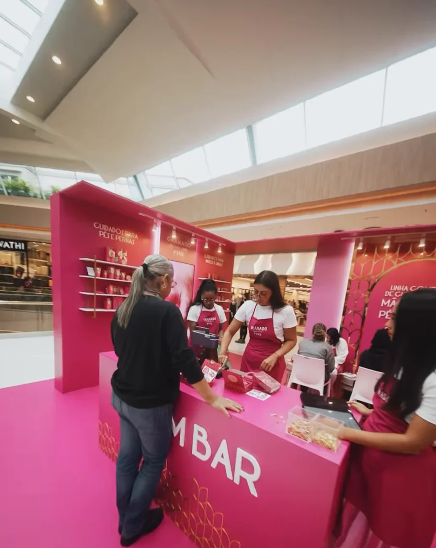 Visão geral de um quiosque pop-up totalmente rosa em um shopping center. Funcionárias de avental rosa e camiseta branca atendem clientes em um balcão onde se lê "BAR".