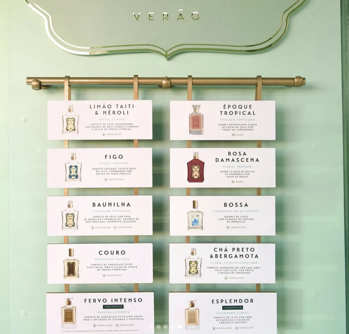 Menu de parede em tom verde-claro sob um letreiro dourado escrito "VERÃO", mostrando placas brancas com nomes de fragrâncias e descrições de sorvetes correspondentes.