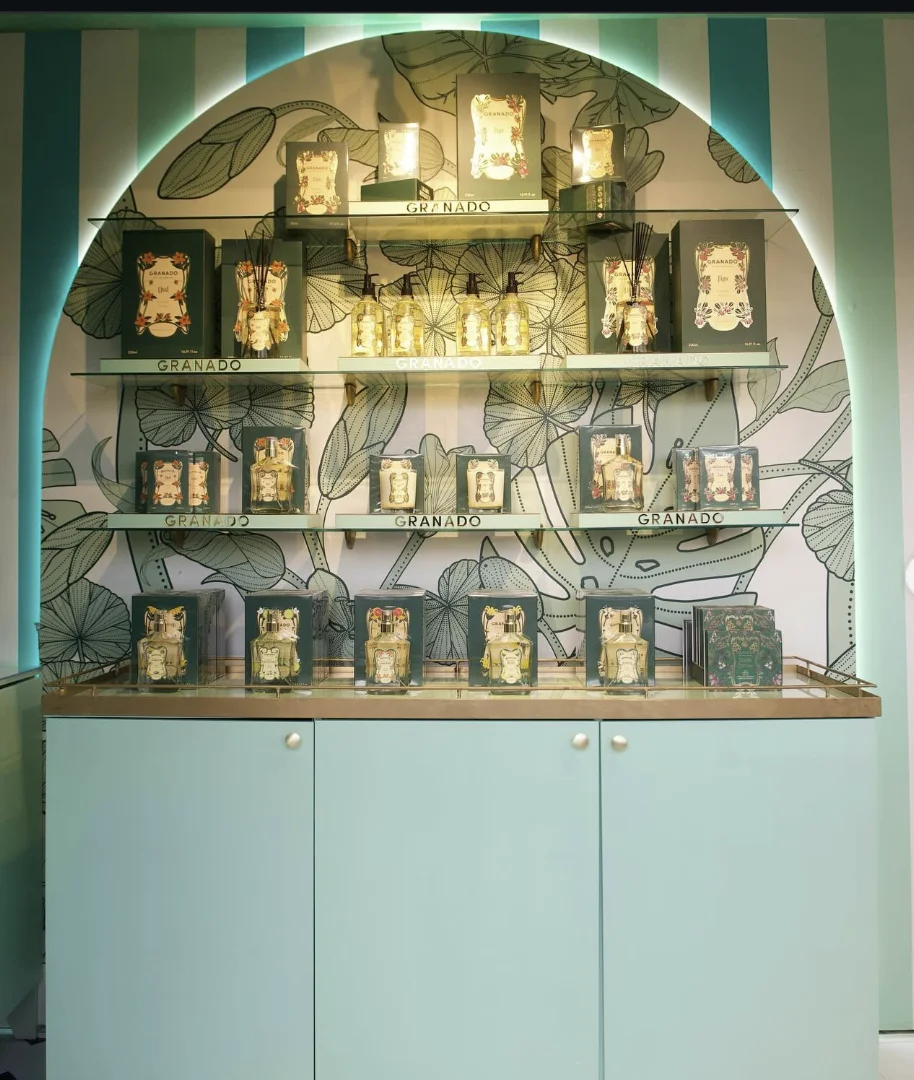 Prateleiras de vidro iluminadas dentro de um nicho em arco, exibindo perfumes, difusores e sabonetes da Granado sobre um balcão verde-claro.