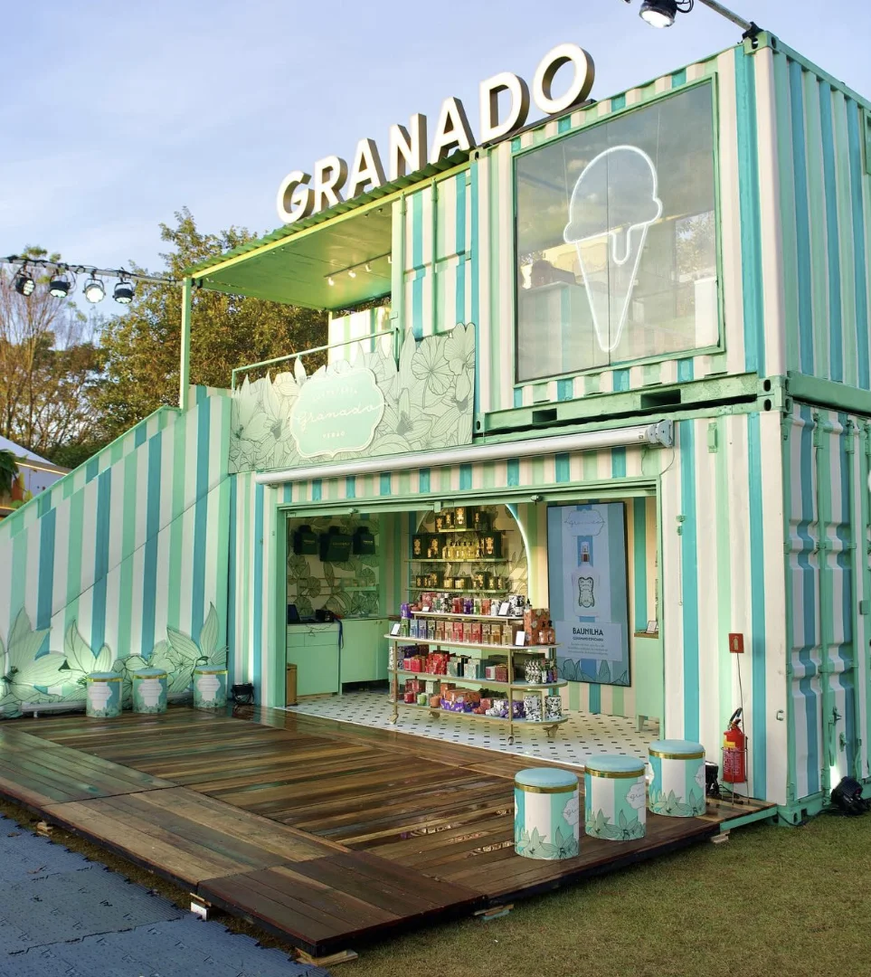 Visão em ângulo diagonal do estande de contêiner listrado da Granado, mostrando o deck de madeira na frente e banquetas decoradas espalhadas pelo gramado.
