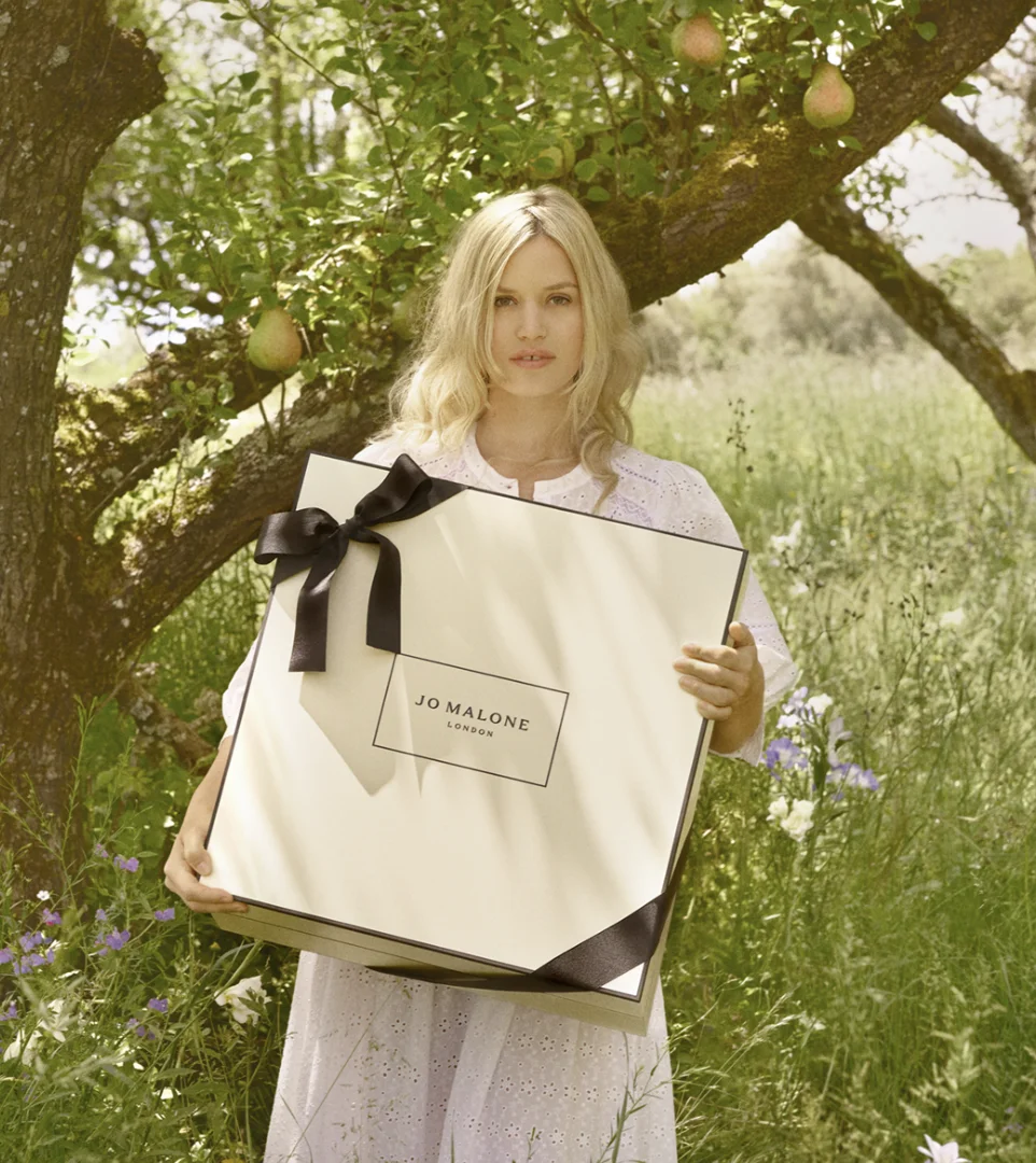 Uma mulher loira com um vestido branco segurando uma caixa de presente grande e creme da marca Jo Malone em um pomar de peras com flores silvestres.