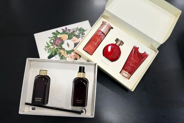 Dois kits de presente da marca O Boticário sobre uma superfície escura. Um kit contém produtos de perfumaria vermelhos em uma caixa clara, e o outro, da linha Casa 214, contém frascos escuros com difusores em uma caixa branca.