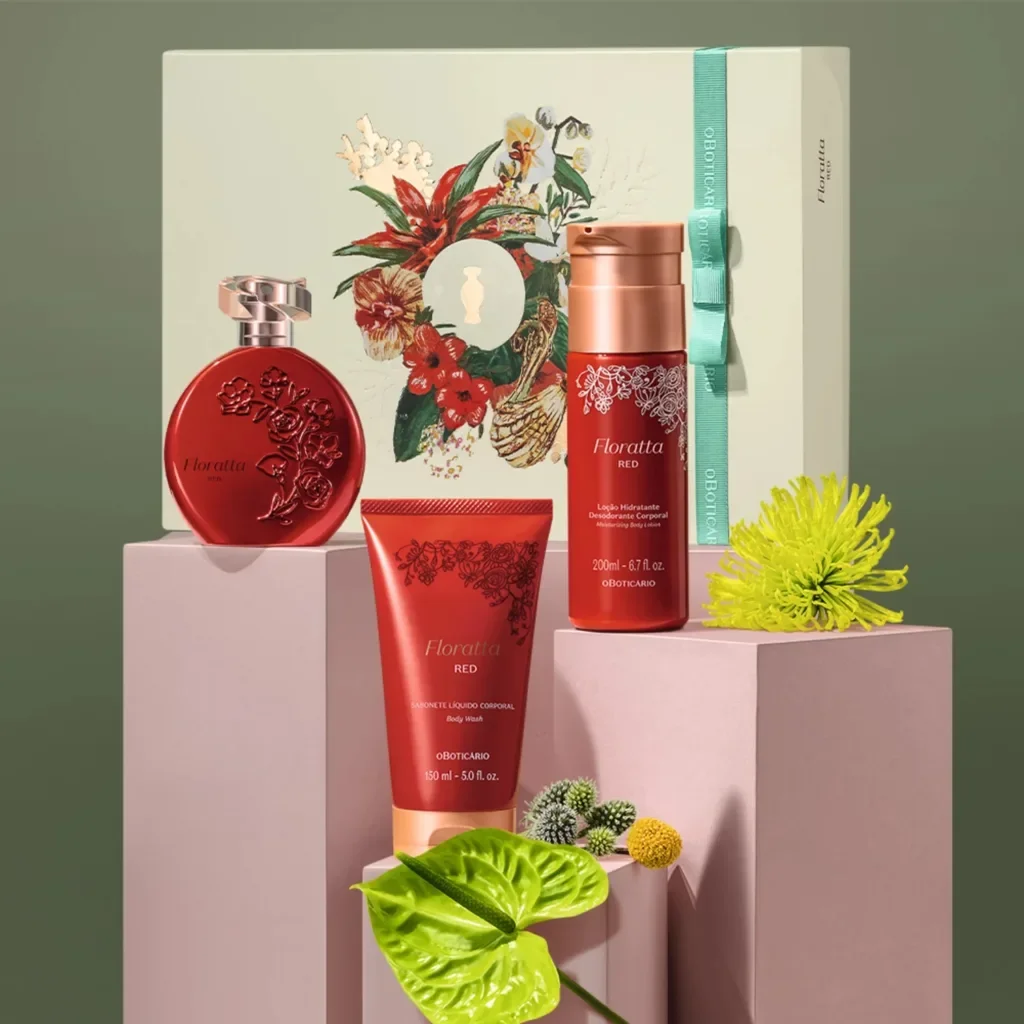 Um arranjo de estúdio com quatro produtos O Boticário Floratta Red e uma caixa de presente floral. A caixa está no fundo em um pedestal rosa claro com uma fita turquesa. À sua frente estão dois perfumes vermelhos redondos e um frasco de spray de loção corporal vermelho. No primeiro plano, uma folha verde grande está sobre um pedestal menor.