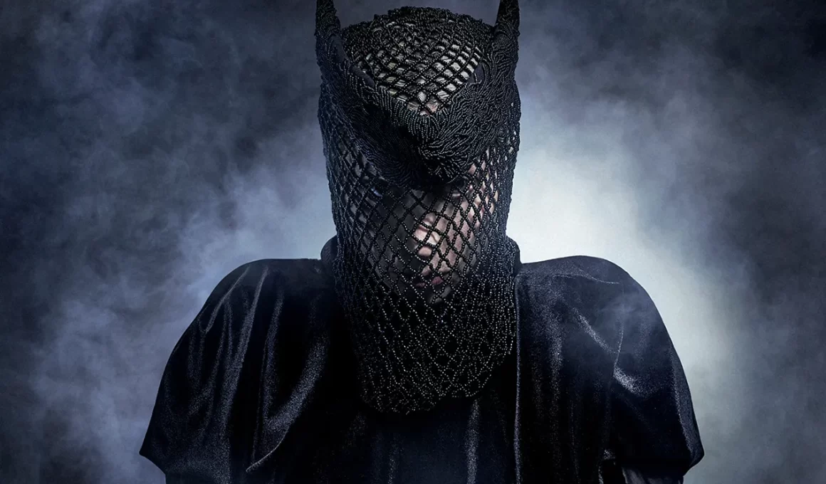 Close-up de Lady Gaga vestindo um look dramático todo preto em meio a uma névoa cinzenta. Ela usa uma máscara de tela preta intrincada que cobre todo o seu rosto e cabeça, revelando vagamente seus olhos, com pequenas pontas que lembram chifres no topo.