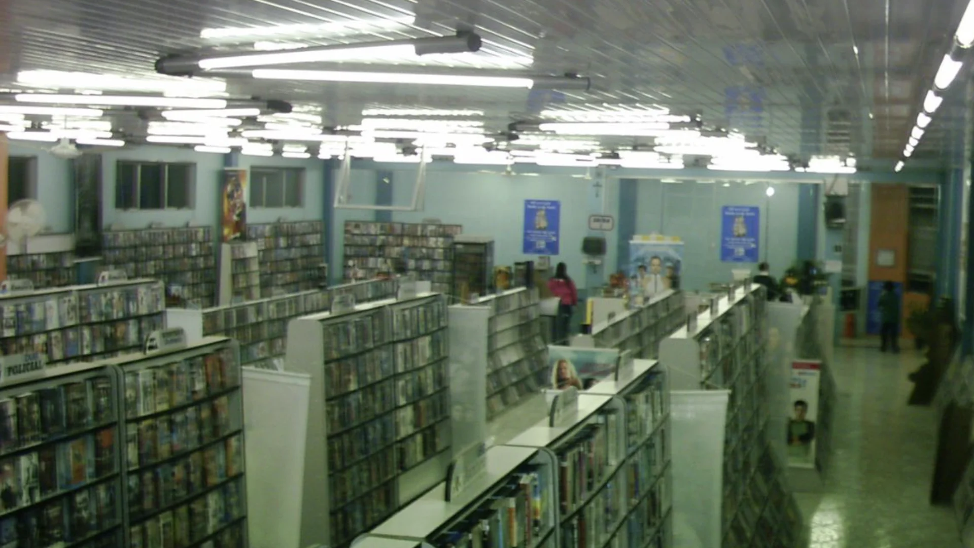 Interior de uma locadora de vídeos iluminada por luzes fluorescentes, com diversas estantes repletas de fitas VHS e DVDs organizados por gênero.