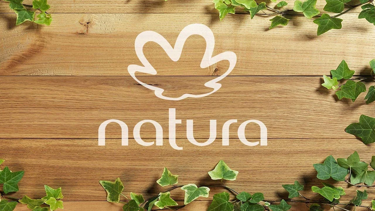 Logotipo da Natura em branco centralizado sobre um fundo de tábuas de madeira clara, emoldurado por ramos de folhas verdes nas bordas superior e inferior.