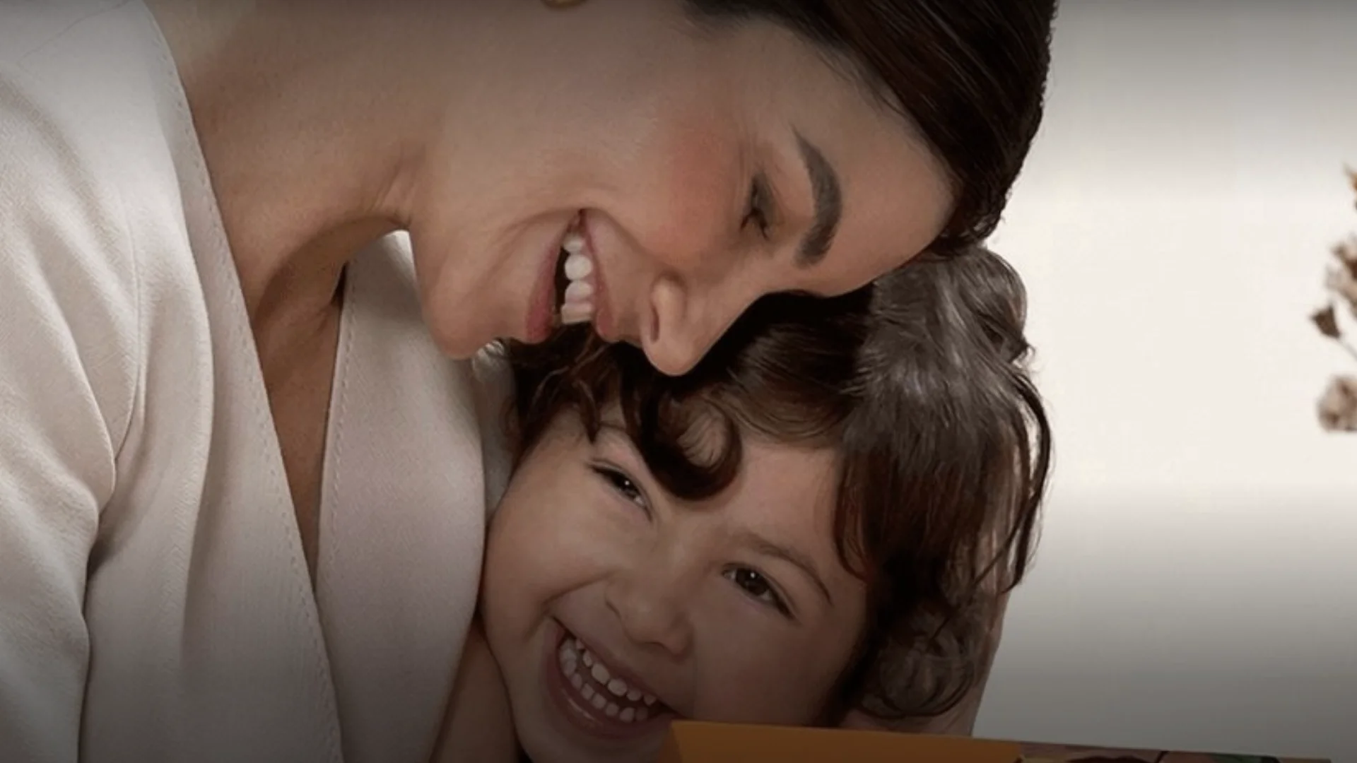 Close de uma mãe sorridente com o rosto encostado no de seu filho pequeno, que dá uma risada alegre.