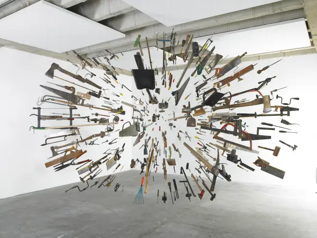 Instalação de arte contemporânea com centenas de ferramentas manuais suspensas por fios invisíveis, formando uma grande esfera flutuante em uma sala branca.