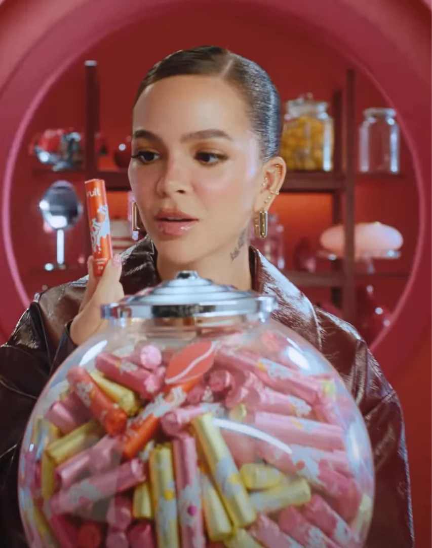 A atriz Mel Maia segura um gloss labial da Vult enquanto olha para uma grande bomboniere de vidro cheia de produtos coloridos em formato de chocolate Baton.