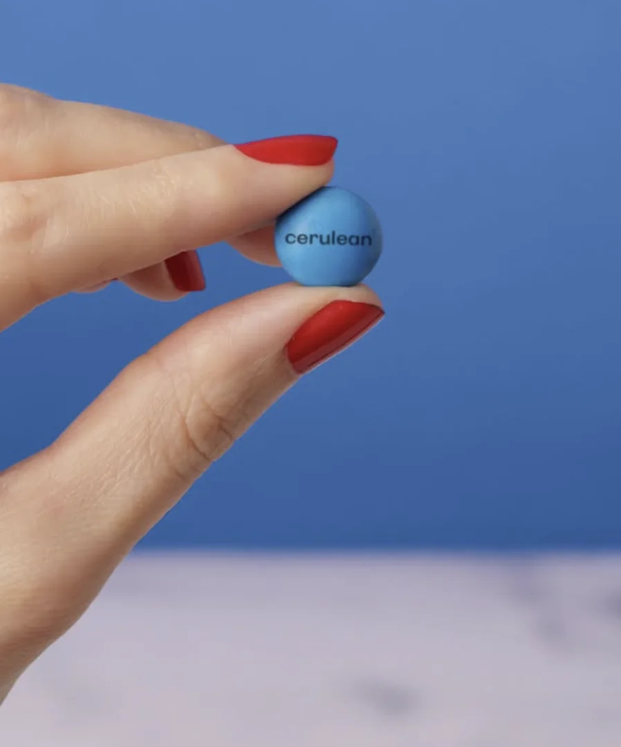 Close-up de uma mão com unhas pintadas de vermelho segurando um único confeito M&M azul. Em vez da letra "m", o confeito tem a palavra "cerulean" escrita em preto.