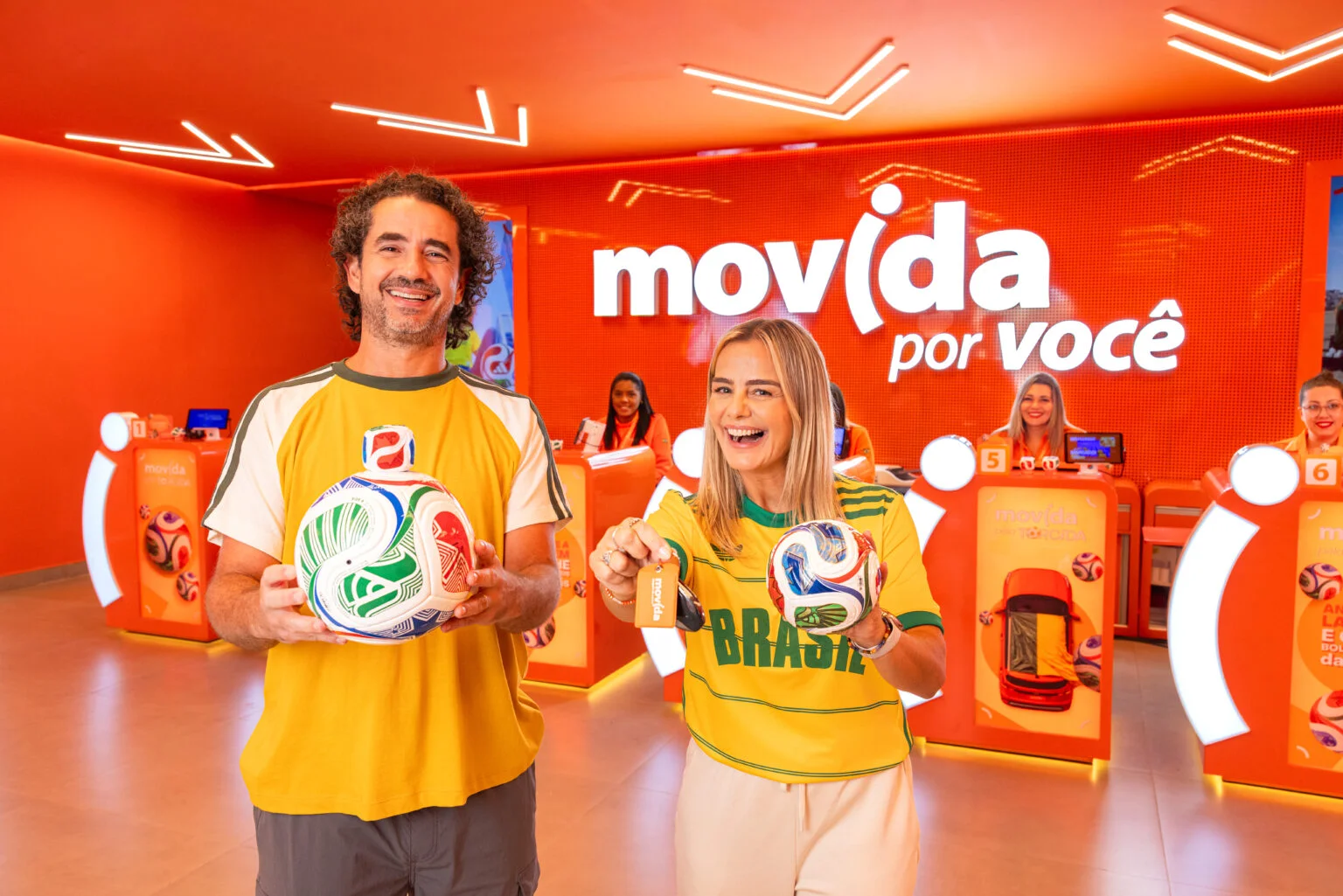 Homem e mulher sorridentes no balcão de atendimento da Movida, segurando bolas de futebol e chaves de carro. O fundo possui um painel laranja vibrante com o logo "Movida por você".
