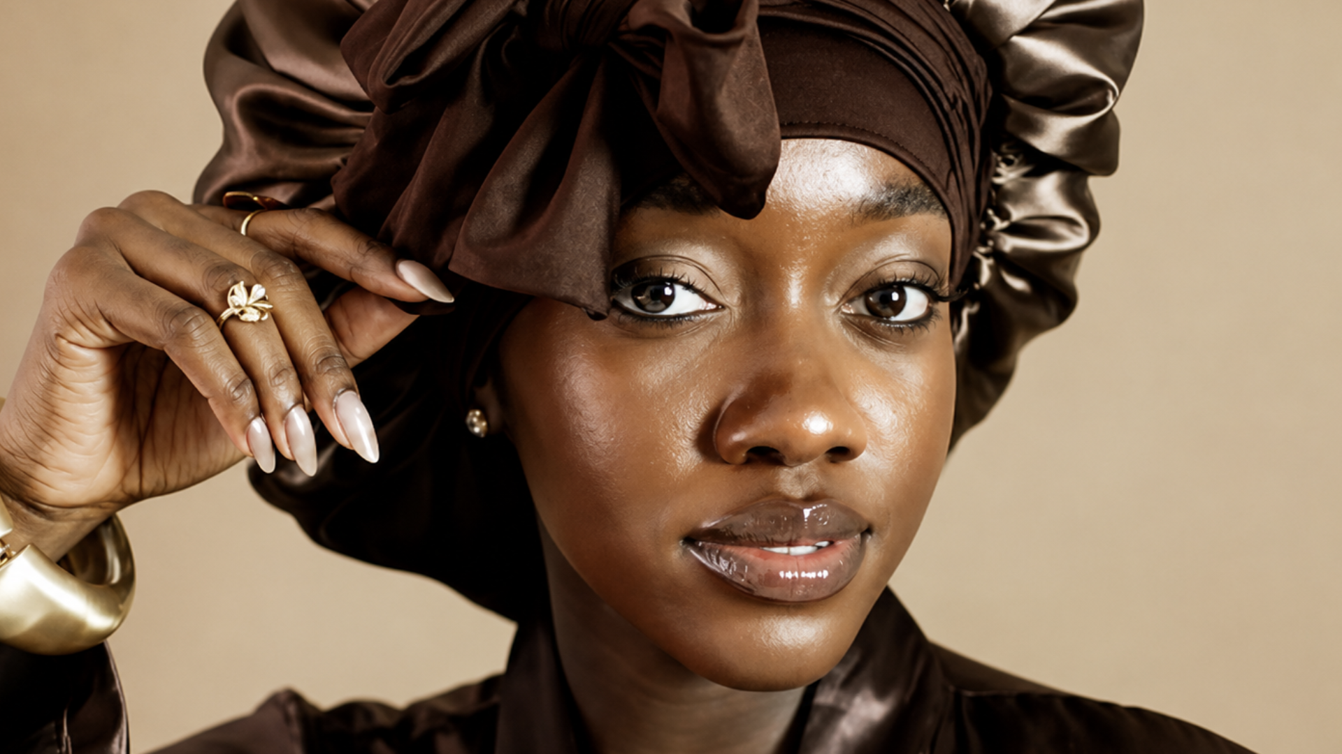 Close-up de uma mulher negra usando uma touca de cetim marrom-escura com um laço frontal volumoso. Ela tem a pele iluminada, lábios com gloss translúcido e exibe anéis dourados e unhas longas pintadas em tom claro enquanto toca delicadamente a lateral da touca.