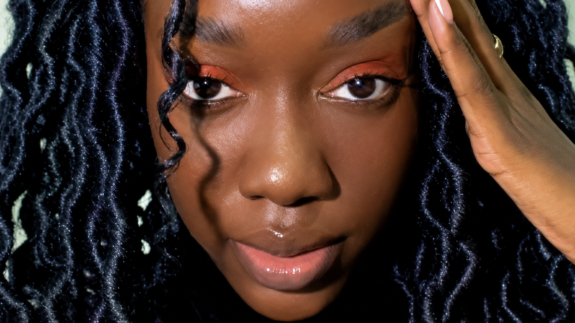 Close-up do rosto de uma jovem mulher negra com cabelos cacheados/tranças escuras. Ela tem uma maquiagem nos olhos em tons quentes de terracota, lábios com brilho labial e posa com a mão esquerda levemente apoiada na têmpora, olhando diretamente para a câmera.