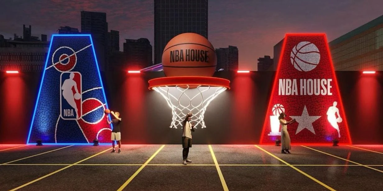 Fachada da NBA House ao anoitecer, apresentando uma grande estrutura central com uma cesta e uma bola de basquete gigante em cima, ladeada por dois painéis trapezoidais iluminados em neon azul e vermelho.