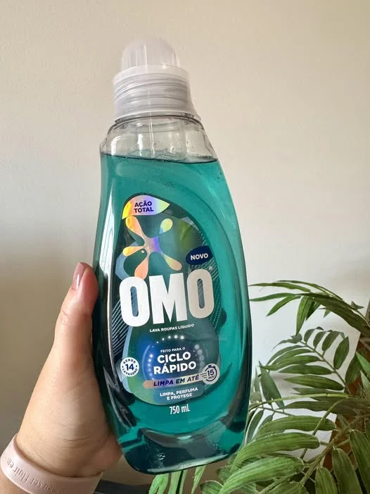 Mão segurando um frasco transparente de 750 ml do lava-roupas líquido OMO Ciclo Rápido, que possui um líquido verde-azulado. Ao fundo, uma parede clara e folhas de uma planta.