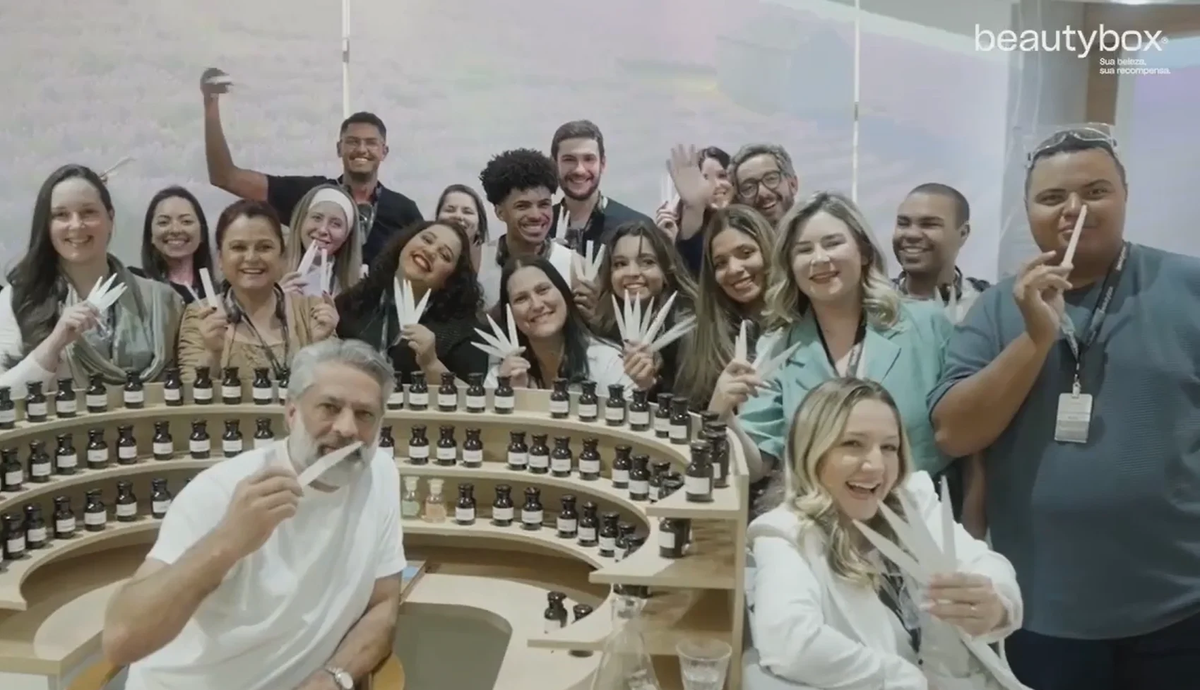 Grupo de cerca de vinte pessoas sorridentes posa para foto segurando fitas olfativas de papel em volta de uma bancada circular com frascos de perfume.