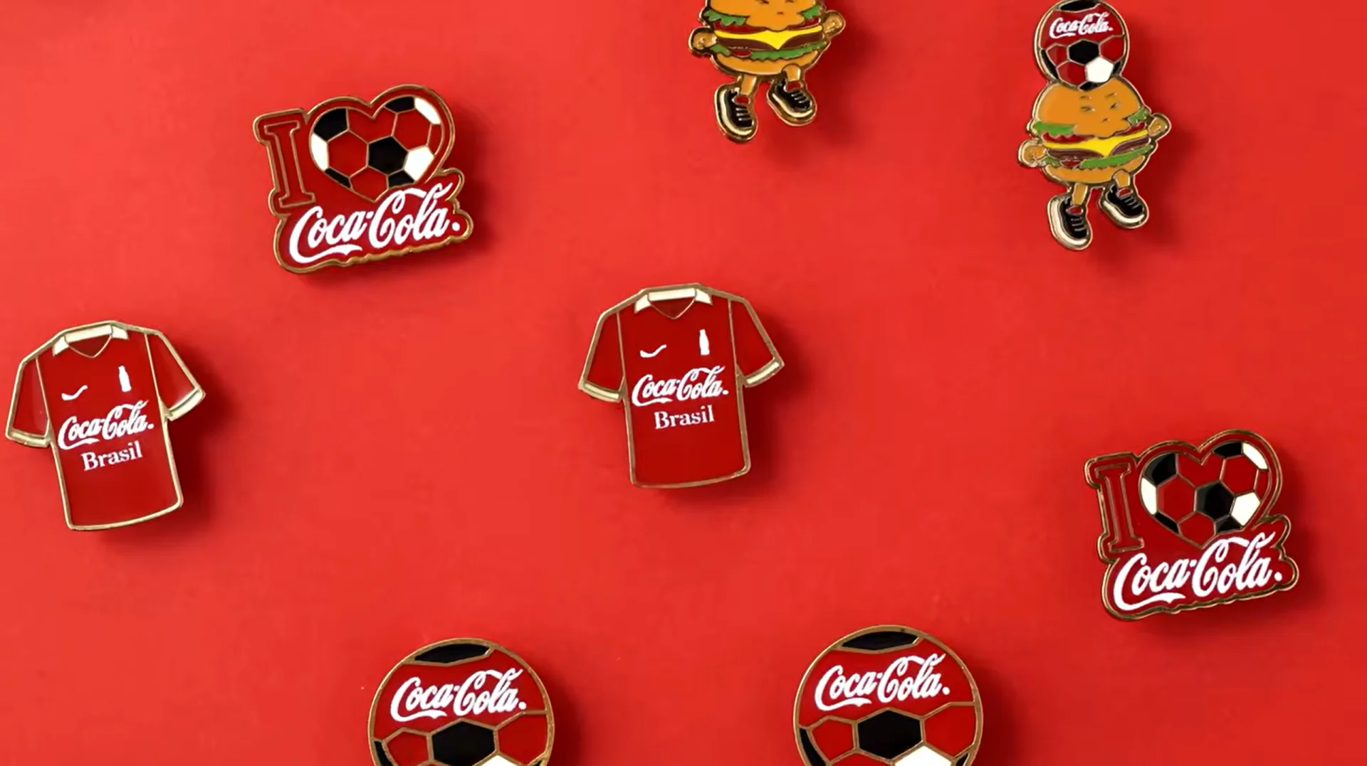 Vários pins colecionáveis da Coca-Cola com tema de futebol espalhados de forma organizada sobre um fundo totalmente vermelho.