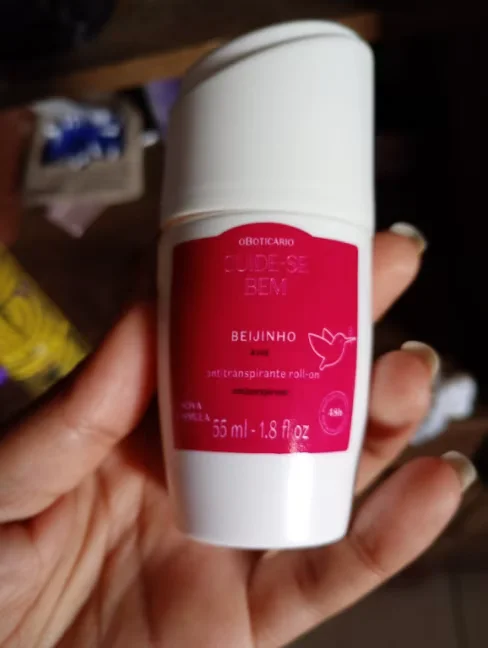 Mão segurando um desodorante antitranspirante roll-on da linha Cuide-se Bem Beijinho, de O Boticário, com embalagem branca e rótulo rosa.
